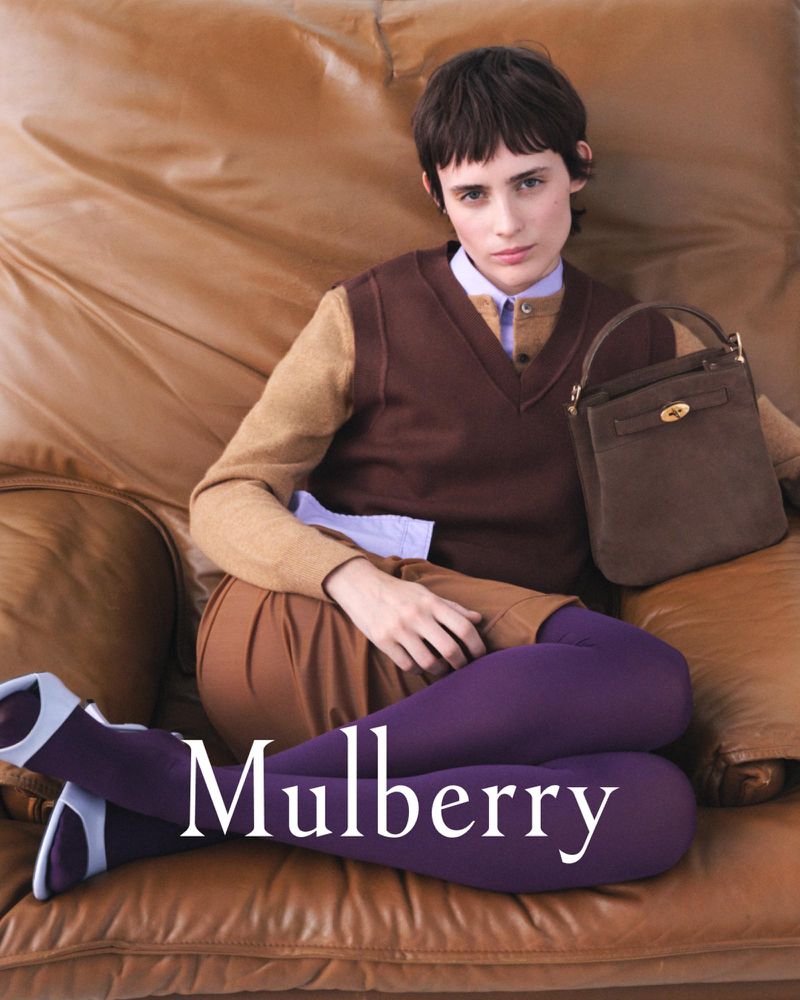 A woman poses on a leather couch holding a Mulberry handbag in an indoor setting, showcasing fashion and style with natural lighting.