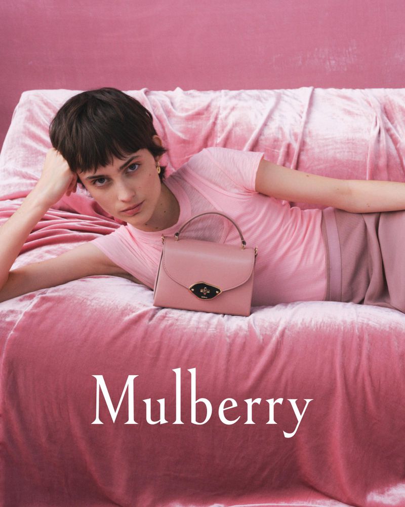 A woman poses while reclining on a pink velvet surface next to a pink leather Mulberry handbag, against a matching pink background.