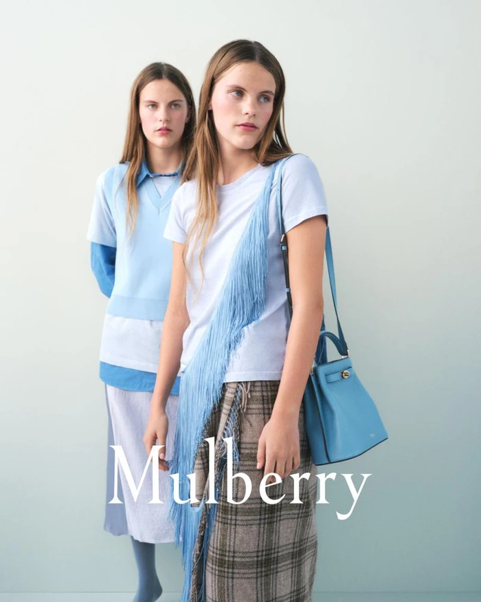 Two female models posing for the Mulberry T2 campaign against a pale gradient backdrop wearing blue apparel and holding a blue handbag
