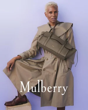 Model with short blonde hair wearing a beige belted trench coat and holding a green leather handbag against a pale purple background.