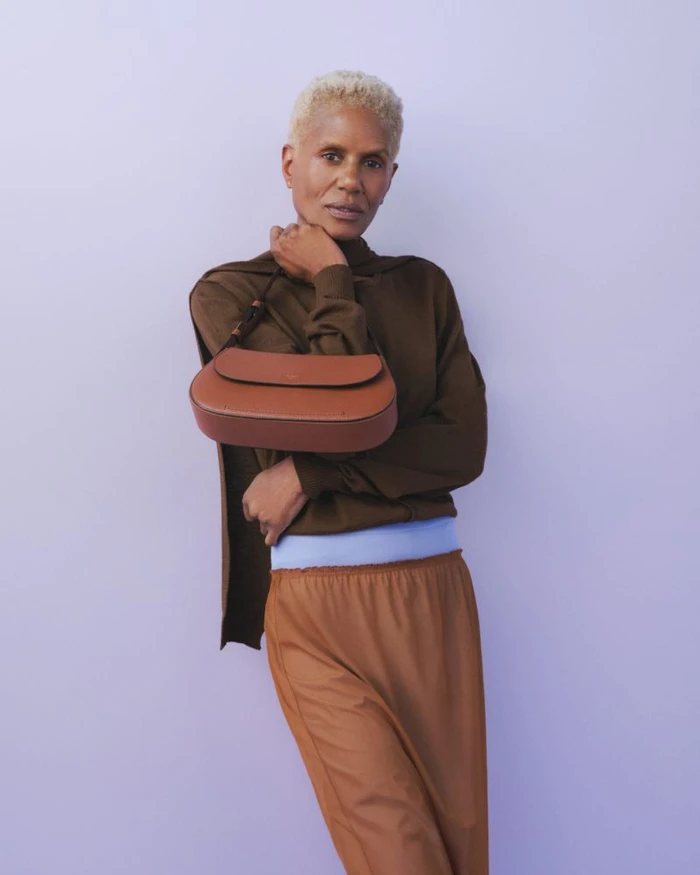 Model with short blonde hair wearing a brown top and skirt holding a brown leather handbag against a pale purple backdrop.