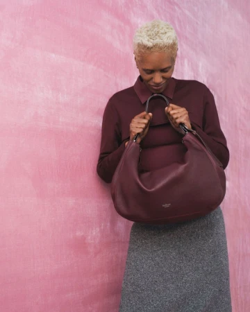 Model with short blonde hair wearing a dark burgundy top and grey skirt holding a large leather handbag against a pink textured backdrop
