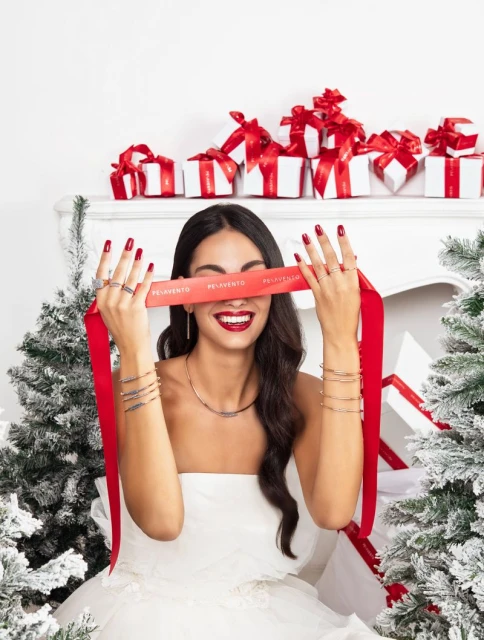 Smiling woman in white dress holds branded ribbon over eyes, adorned with jewelry; gift boxes and Christmas trees create festive backdrop.