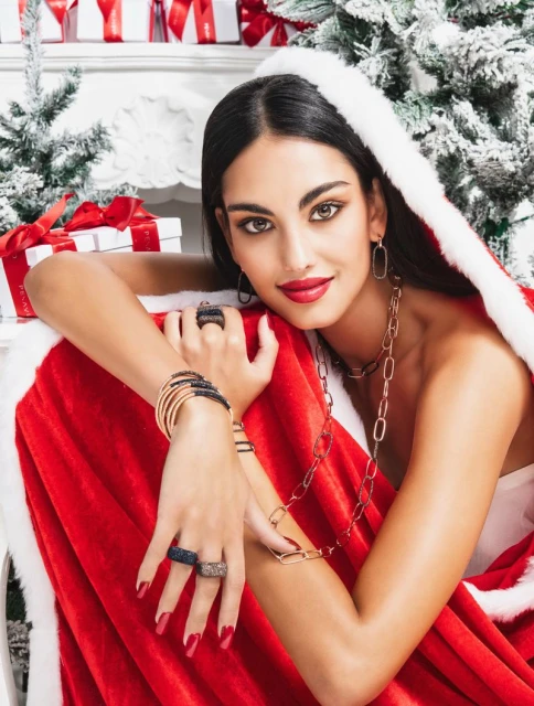 Woman with jewelry wearing a Santa-style cape, posing in front of a Christmas tree and gift boxes. Beauty and fashion advertisement.