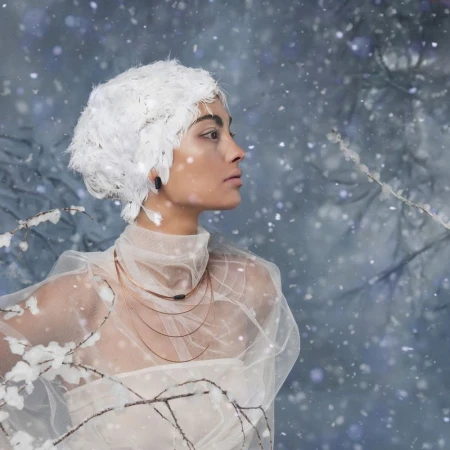 Profile shot of a woman in a white feather headdress, tulle top, and layered necklaces against a snowy blue background with branches.