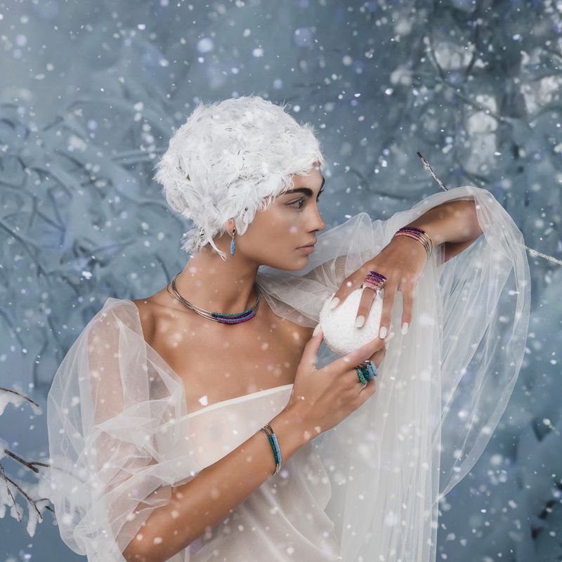 A woman in winter-themed jewelry and a white head covering holds a snowball in an indoor, snowy setting for a beauty or fashion shoot.