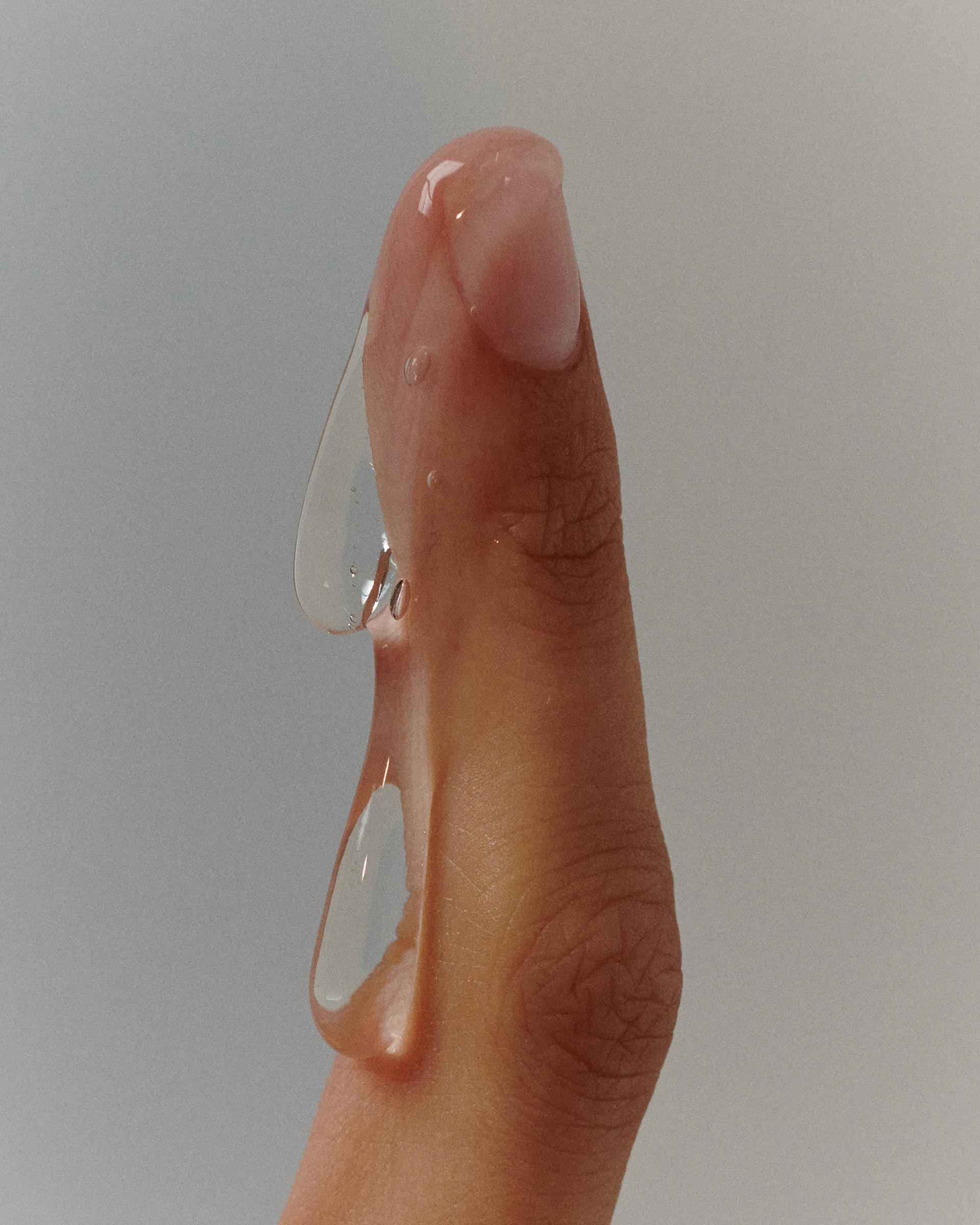 Close-up of a finger with clear, viscous liquid dripping down, showcasing texture, clarity, and skin detail against a muted background.