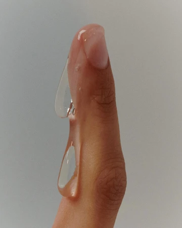 Close-up of a finger with clear, viscous liquid dripping down, showcasing texture, clarity, and skin detail against a muted background.