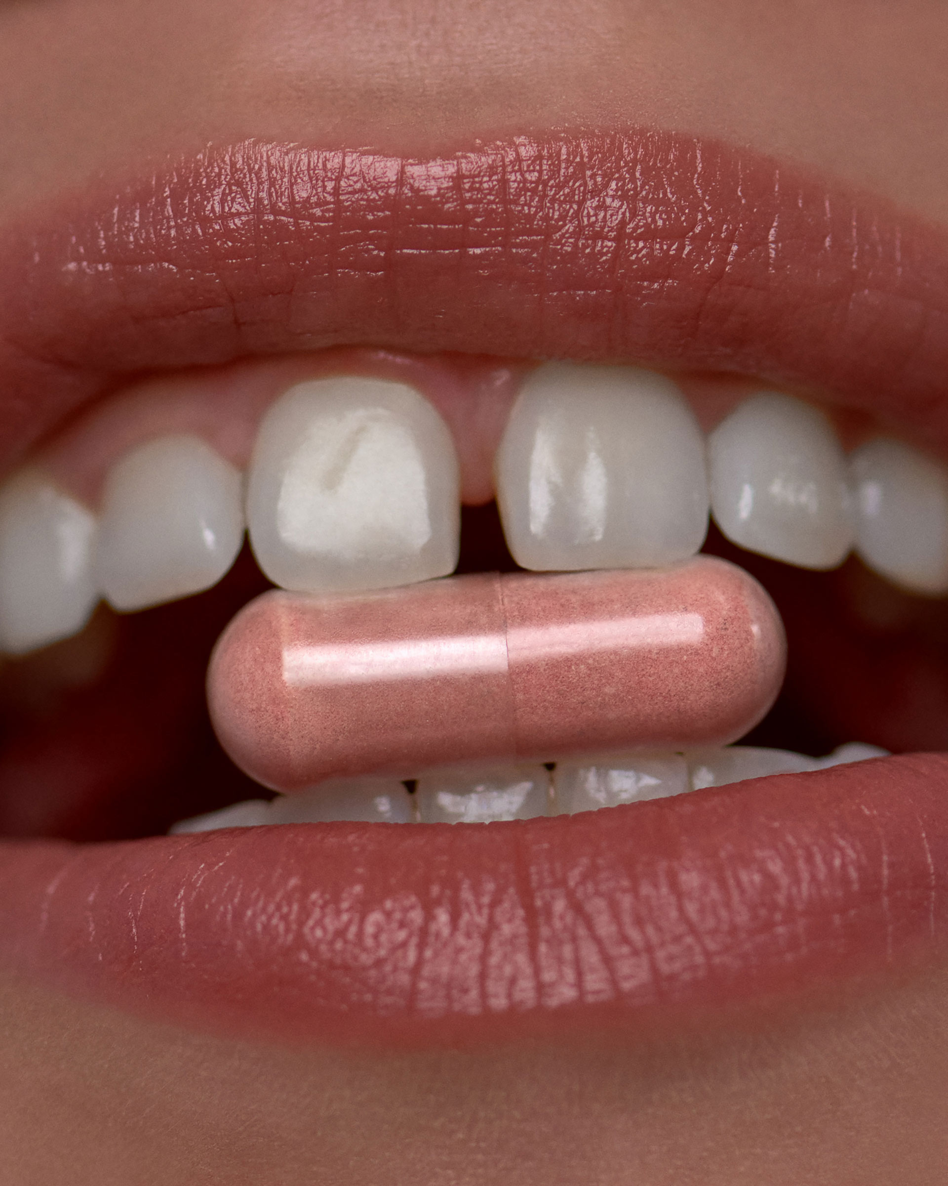 Close-up shot of a pink capsule held between a person's white teeth and pink lips, set against a blurred background.
