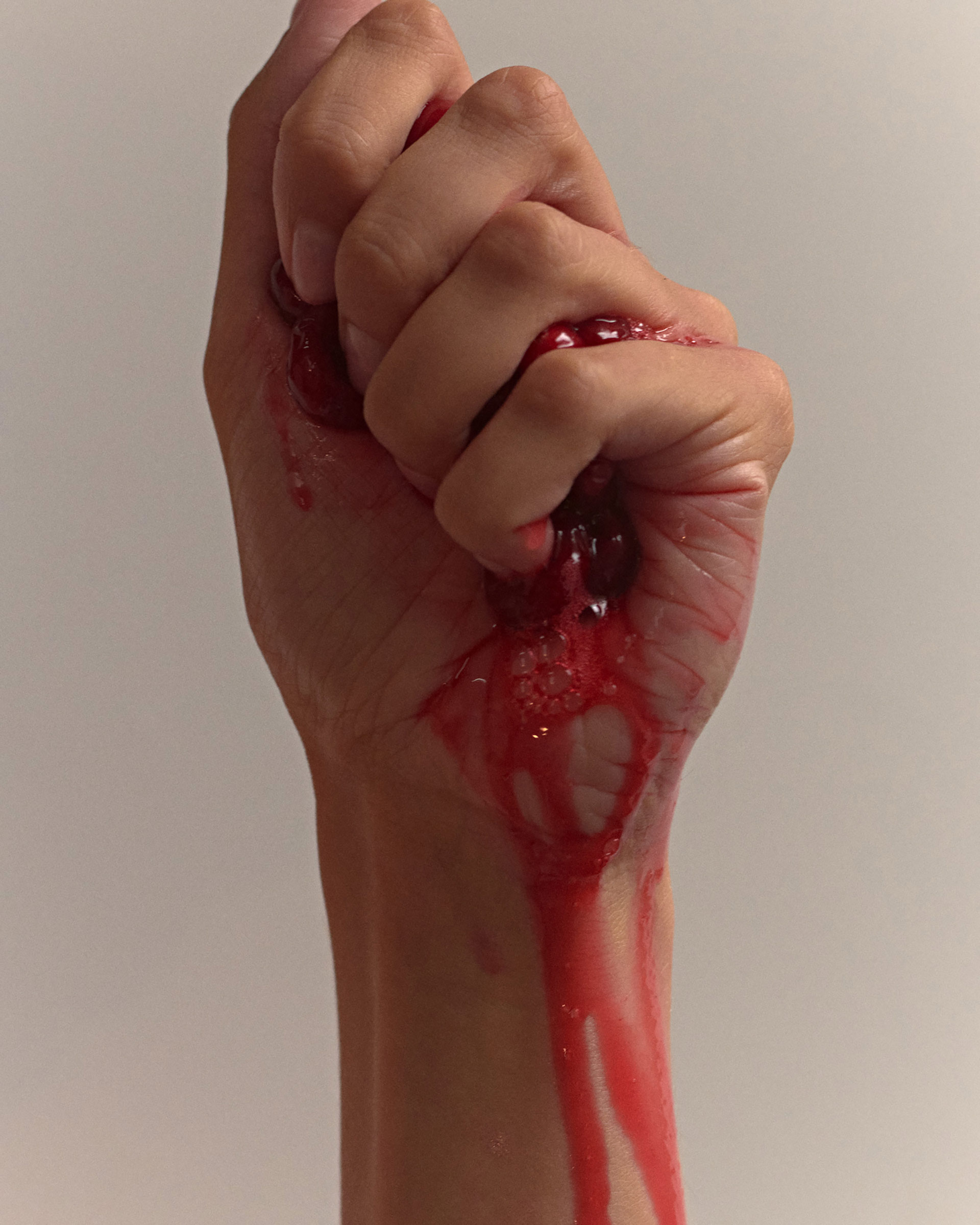 Close-up shot of an adult's arm and hand tightly clenched, covered in dripping, bright red liquid against a plain, neutral background.