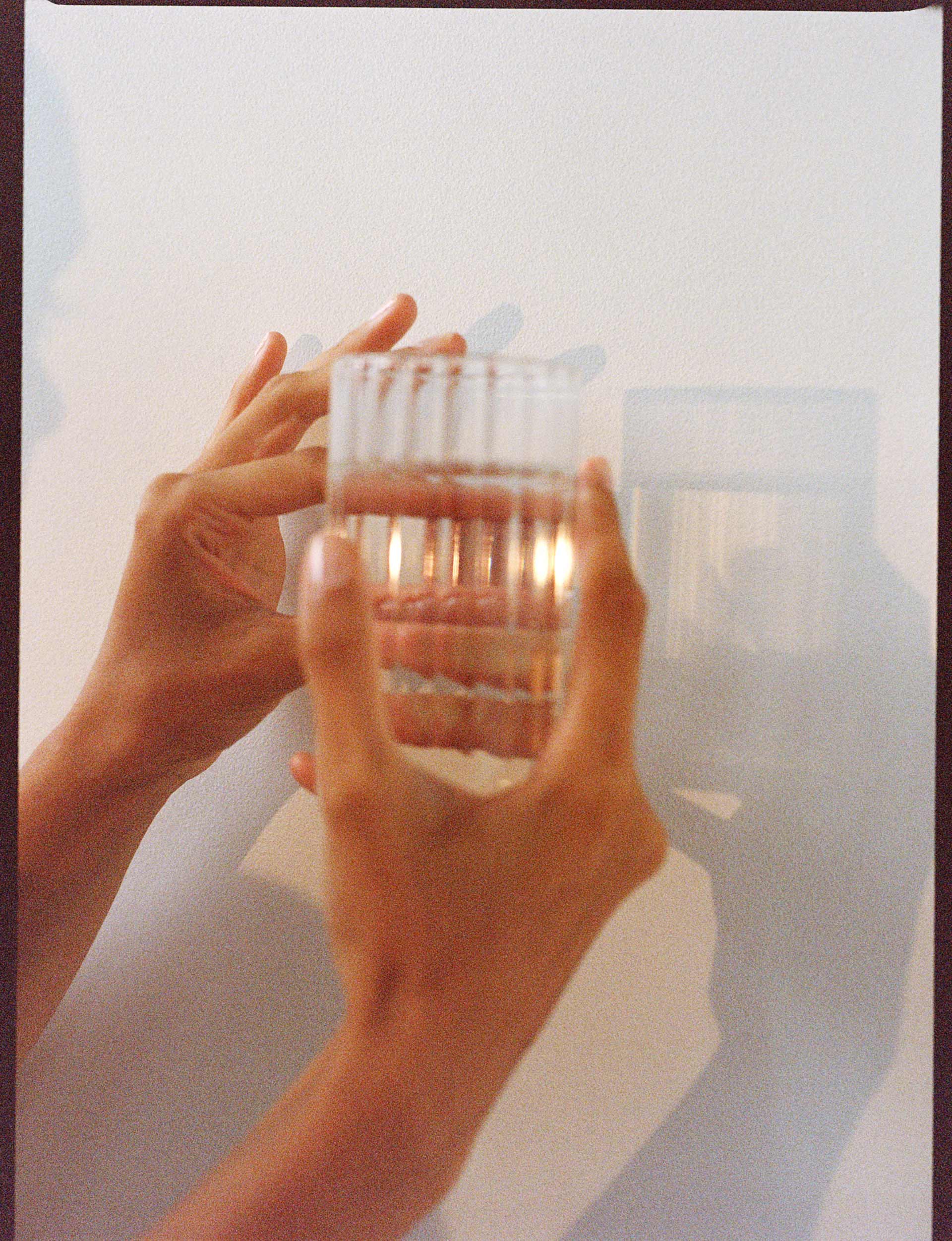 A clear glass of water is held in a pair of hands against a white wall creating a shadow of the glass.