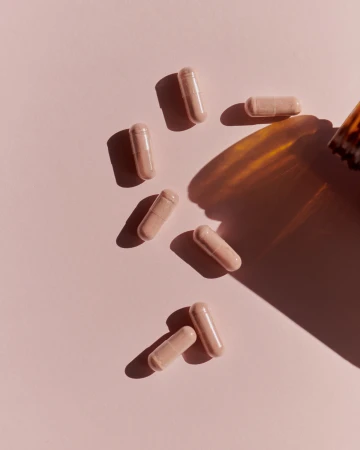 Several light pink capsule pills are scattered on a pastel pink surface, illuminated by a warm, directional light source casting shadows.