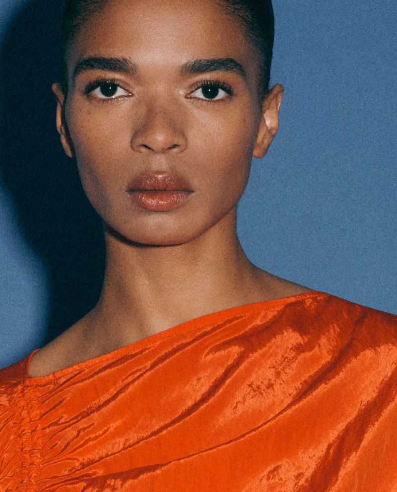 Portrait of a black female adult in a draped orange dress against a plain blue background in a fashion photography style, medium shot.