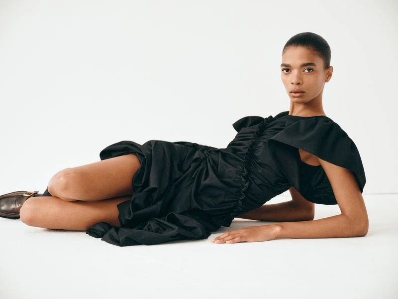 Fashion shot of a woman lying on a white background wearing a black ruffled dress with brown shoes looking directly at the camera.