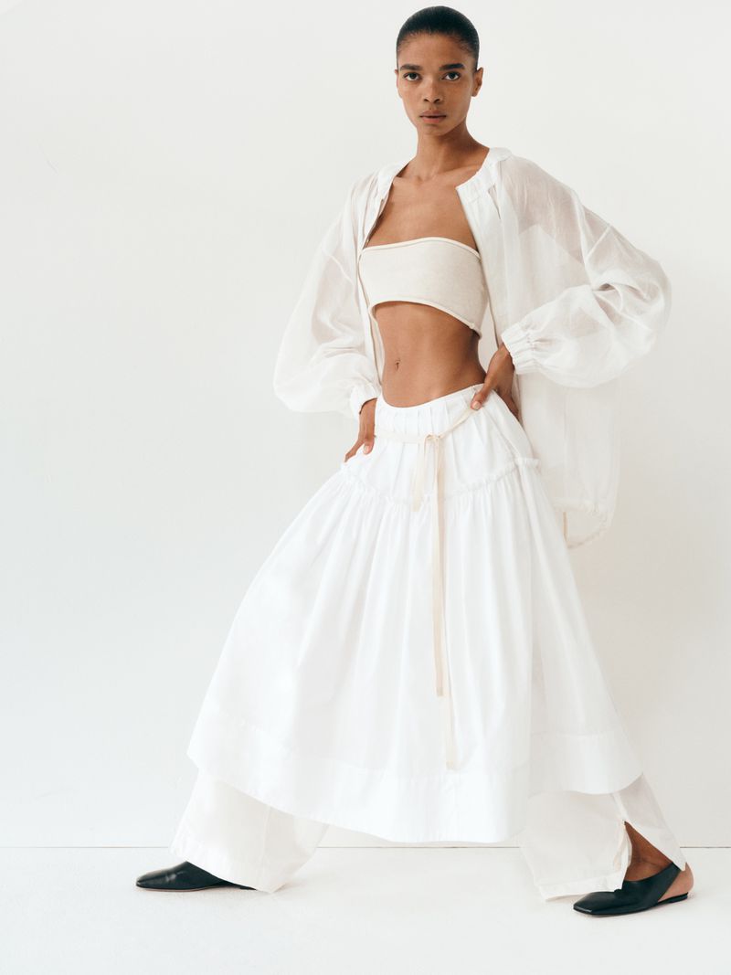Fashion model poses in studio wearing white tube top, sheer oversized jacket, voluminous skirt and pants with black ballet flats.
