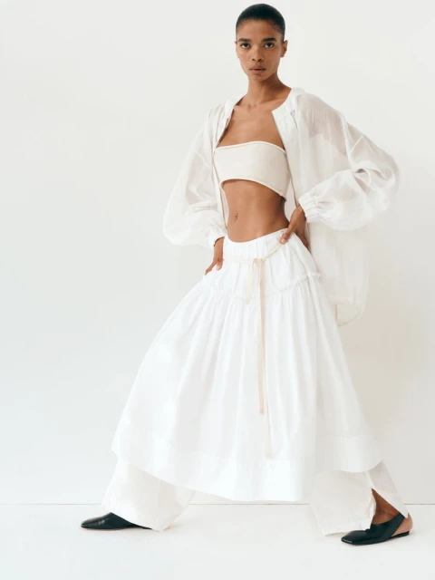 Fashion model poses in studio wearing white tube top, sheer oversized jacket, voluminous skirt and pants with black ballet flats.