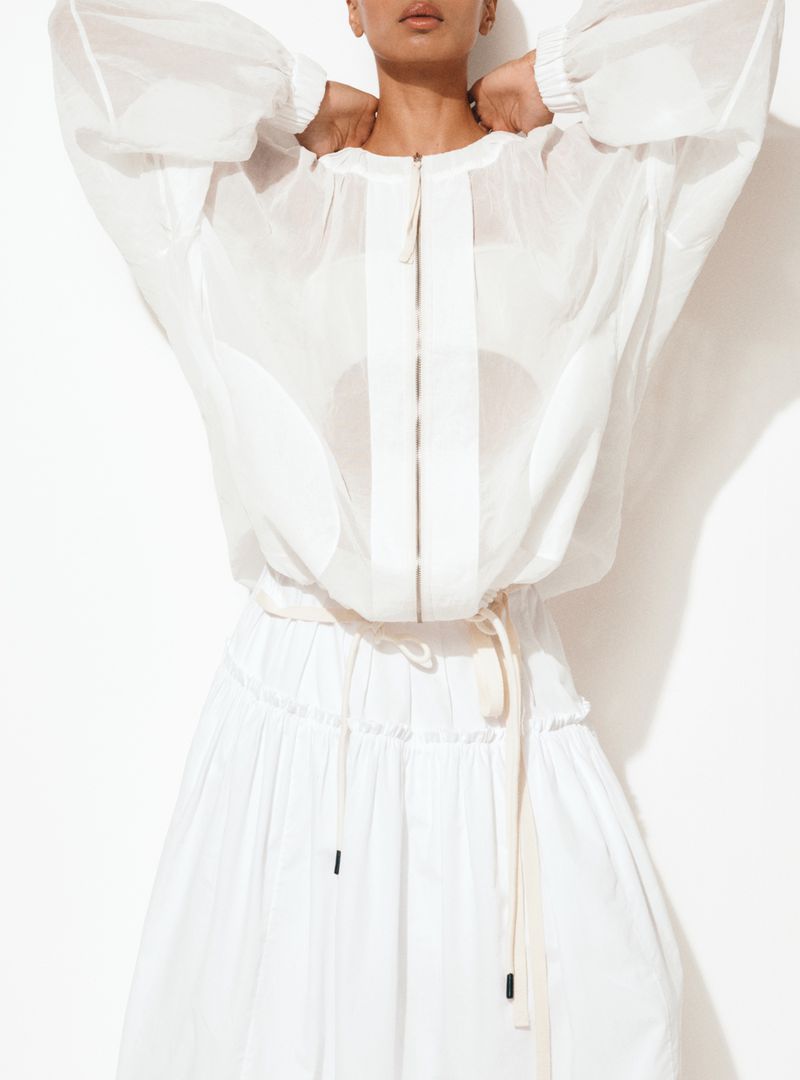 A light-skinned woman is posing in a studio wearing a white, semi-sheer, zip-front jacket and a white, tiered skirt against a white background.