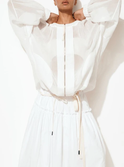 A light-skinned woman is posing in a studio wearing a white, semi-sheer, zip-front jacket and a white, tiered skirt against a white background.