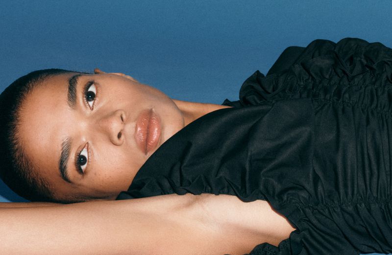 Close-up fashion shot of an adult female with dark hair, wearing a black ruffled top, lying down on a blue backdrop in a photo studio.