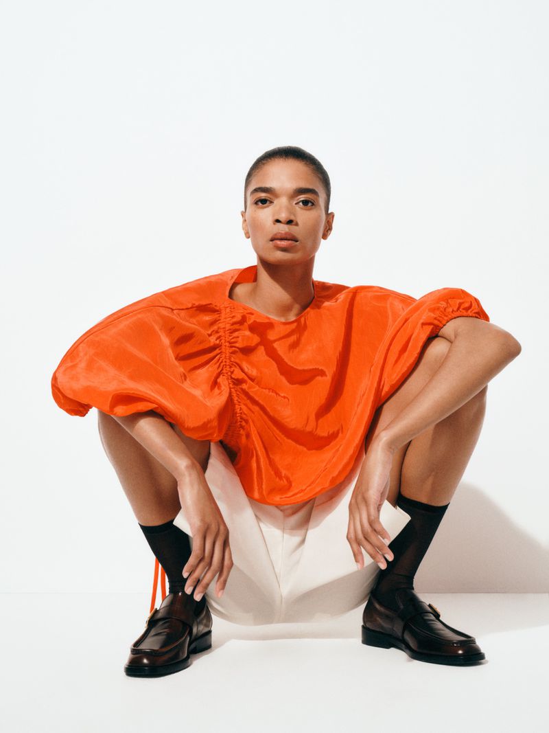 A woman in an orange top and shorts poses while crouching in a studio setting with neutral white background, fashionable and confident.