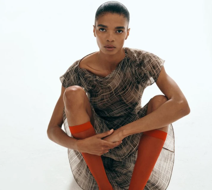 Woman in plaid dress and orange socks sits in front of a white background in a fashion studio portrait with medium shot.