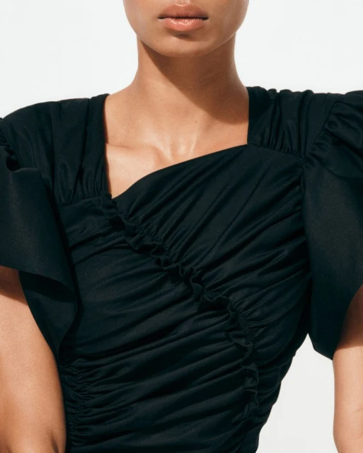 Close-up studio shot of a fair-skinned adult woman with neutral expression wearing a black short sleeve dress against a white background.