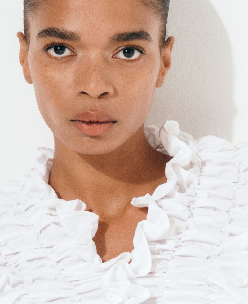 Close-up of a woman with tan skin and dark hair looking directly at the camera, wearing a white ruffled top against a white backdrop.