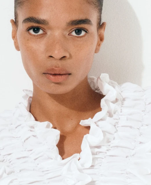 Close-up of a woman with tan skin and dark hair looking directly at the camera, wearing a white ruffled top against a white backdrop.