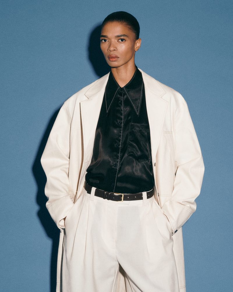 An androgynous person models a cream-colored trench coat, matching trousers, and a black collared shirt against a solid blue backdrop.