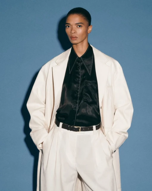An androgynous person models a cream-colored trench coat, matching trousers, and a black collared shirt against a solid blue backdrop.