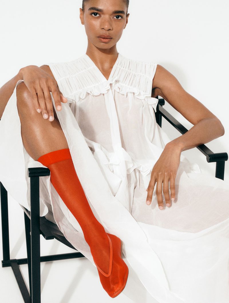 Model seated in a black chair wearing a white sleeveless blouse and vibrant red sheer socks against a white studio wall