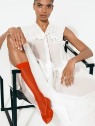 Model seated in a black chair wearing a white sleeveless blouse and vibrant red sheer socks against a white studio wall