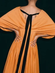 Model seen from behind wearing an orange dress with black back straps against a dark background