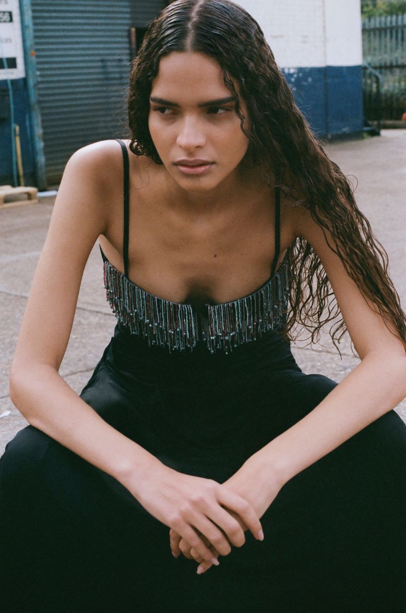 A brunette female model with long wavy hair sits outdoors wearing a spaghetti-strap black top with beaded fringe detailing and black pants.