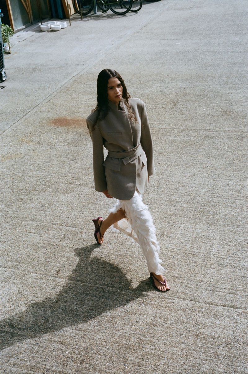 Woman walks across the concrete in a taupe belted jacket with white feathered pants and sandals, captured from above in natural light.