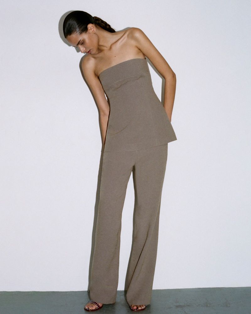 Fashion shoot of a woman in a taupe strapless top and matching trousers in a studio, lit from above, against a stark white background.