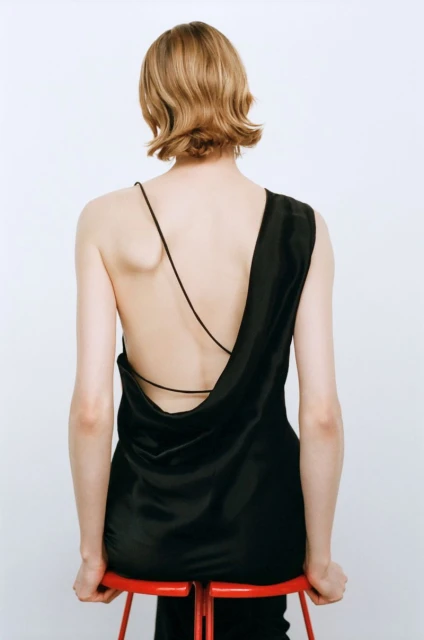 Back view of a woman in a black dress sitting on a red stool against a white background in a fashion forward studio shot.