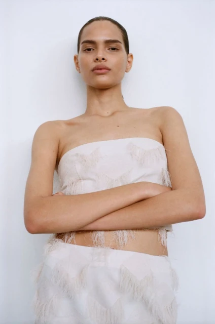 Woman poses in off-white fringe crop top and matching skirt against white backdrop, arms crossed, medium shot, fashion editorial.