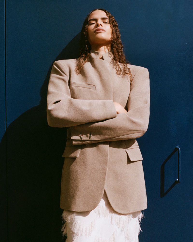 Model with long curly hair wearing a structured beige jacket standing against a deep blue wall for Paris Georgia SS 25.