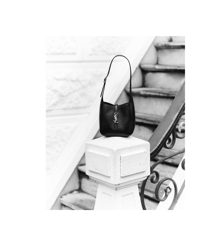 Black Saint Laurent bag on a white column against a grayscale staircase. Minimalist fashion, product shot for handbag advertisement.