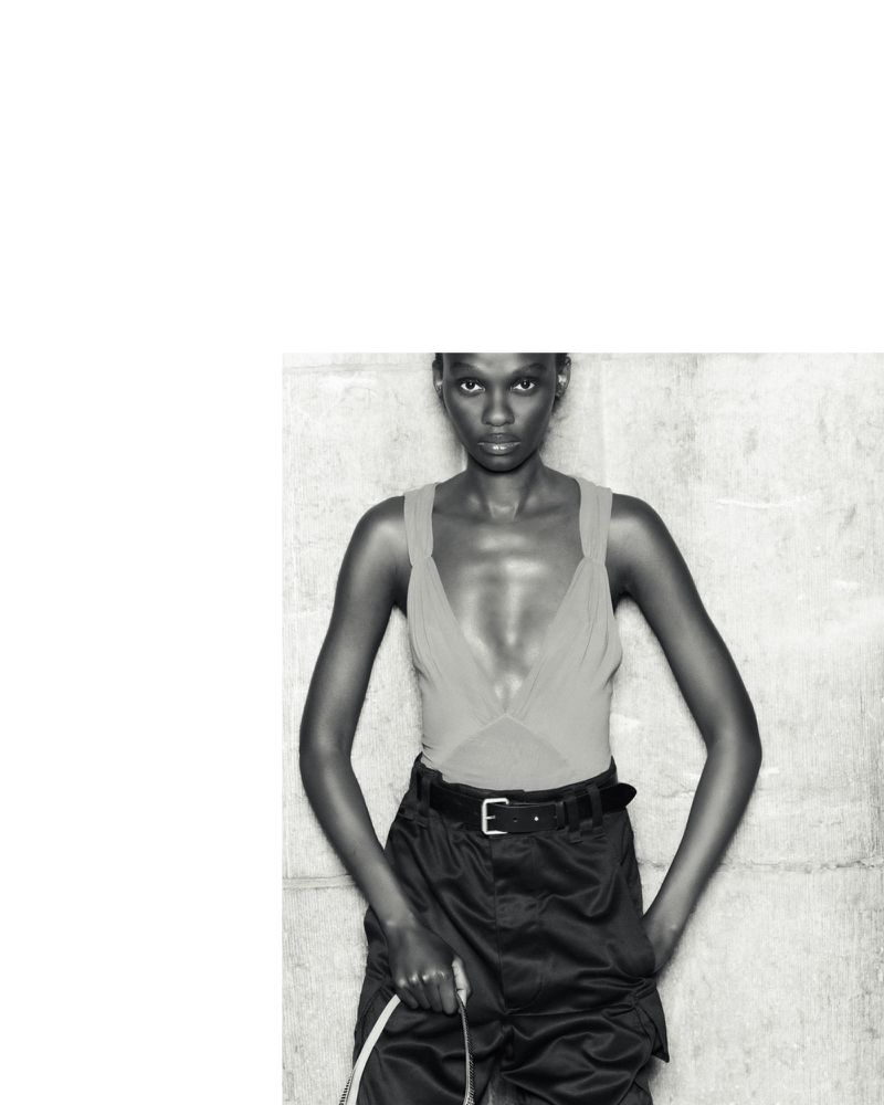 Fashion shot of a Black woman in a sleeveless top and cargo pants with a belt, holding a white cable, against a textured wall.