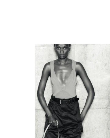 Fashion shot of a Black woman in a sleeveless top and cargo pants with a belt, holding a white cable, against a textured wall.