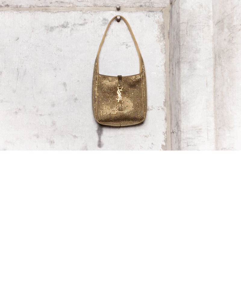 Gold YSL handbag on a silver hook against a textured light gray wall, photographed indoors with a studio setting for a fashion product shot.