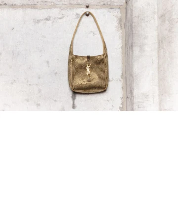 Gold crystal-embossed shoulder bag with metallic YSL hardware hanging on a distressed concrete wall surface.