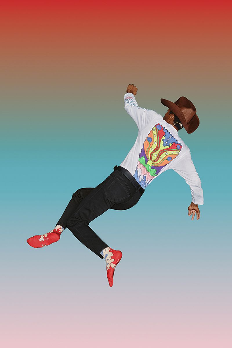 Black adult male in mid-air wearing sunglasses, cowboy hat, graphic tee, and red shoes against a gradient red and blue background.