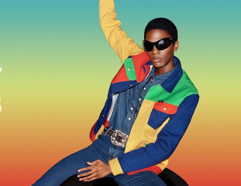 Portrait of a young Black adult wearing colorful patchwork jacket and sunglasses, against a vibrant gradient backdrop, posing dynamically.