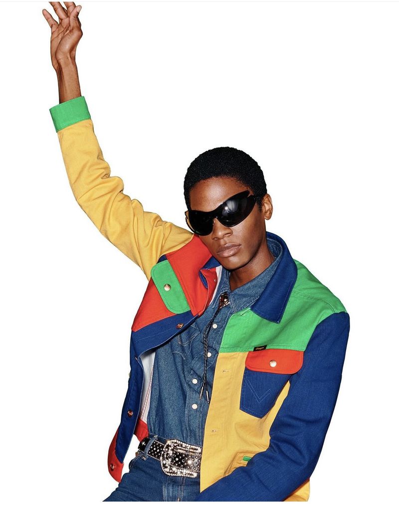 Adult male fashion model in multicolored jacket with denim shirt, ornamental belt, and dark glasses posing against a white backdrop.