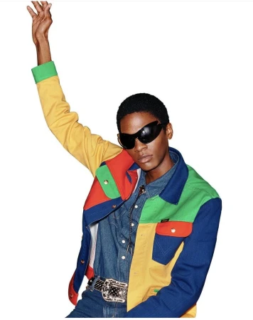 Model wearing a colorful patchwork jacket and dark sunglasses with one arm raised against a plain white background