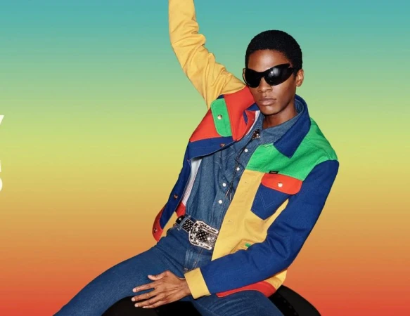 Model wearing dark sunglasses and colourful panelled jacket with denim shirt and jeans against gradient background
