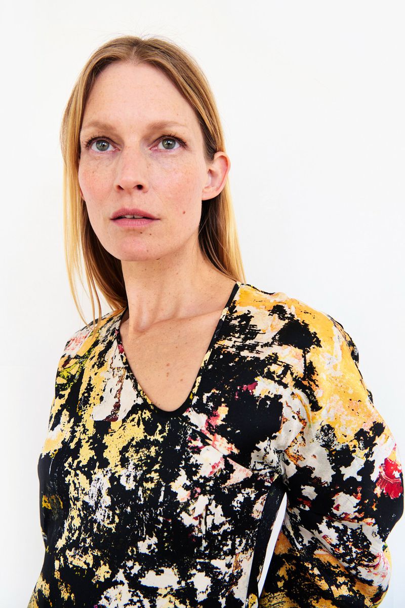 Portrait of an adult woman with long blonde hair, wearing an abstract patterned blouse, against a plain white background in an indoor setting.